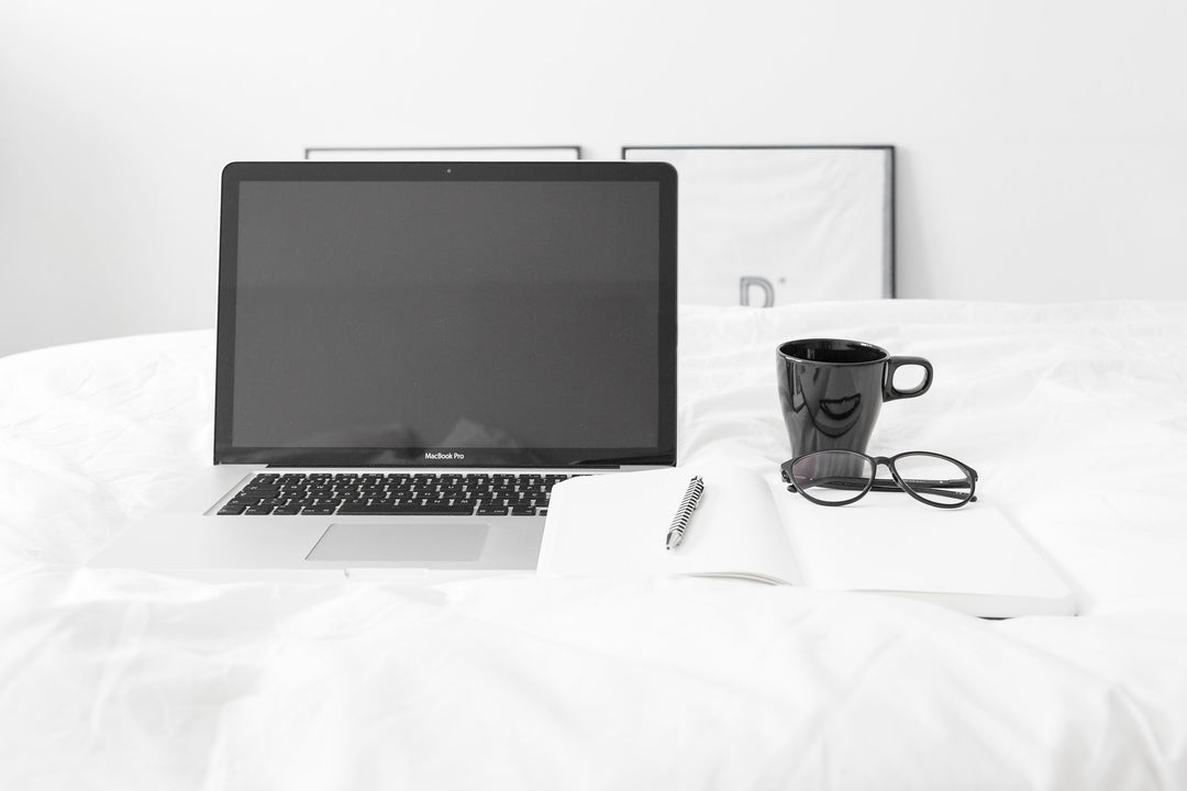 Laptop, notepad, glasses, and coffee mug sitting on a white bedspread