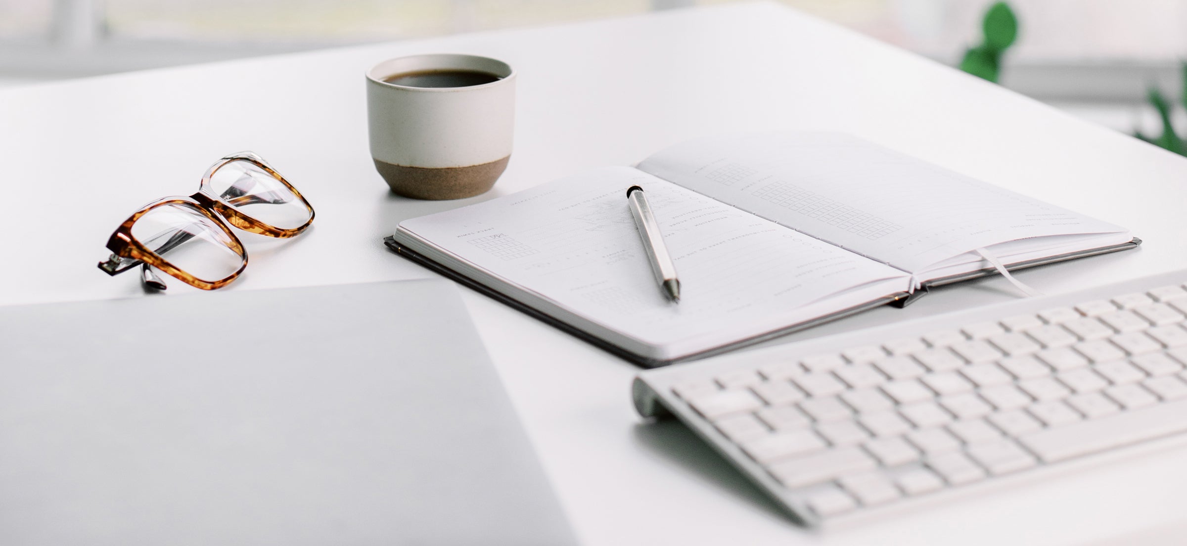 desk with open notebook, coffee, pen, mac keyboard, and glasses